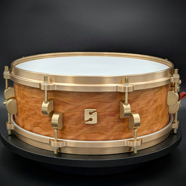 A 5" x 14" North American Quilted Cherry snare drum with brass hardware and a white drum head, displayed on a black background.
